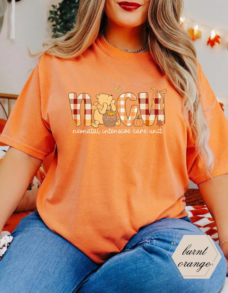 May include: Burnt orange t-shirt with the letters NICU in a plaid pattern, featuring a cartoon bear with a honey pot and a pumpkin. The text "neonatal intensive care unit" is below the letters. The shirt is a burnt orange color.
