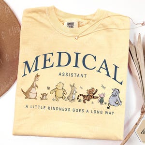 May include: Pale yellow t-shirt featuring the words "MEDICAL ASSISTANT" in navy blue. Below the text are cartoon characters. The shirt also has the phrase "A LITTLE KINDNESS GOES A LONG WAY".
