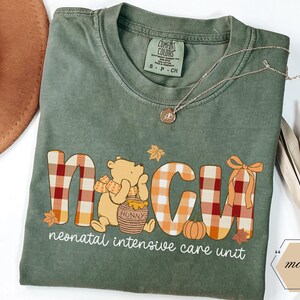 May include: Green Comfort Colors t-shirt with a Winnie the Pooh-themed design. The design includes the letters "NICU" in a plaid pattern, a bear, a pumpkin, and the words "neonatal intensive care unit". A gold necklace is draped across the shirt.