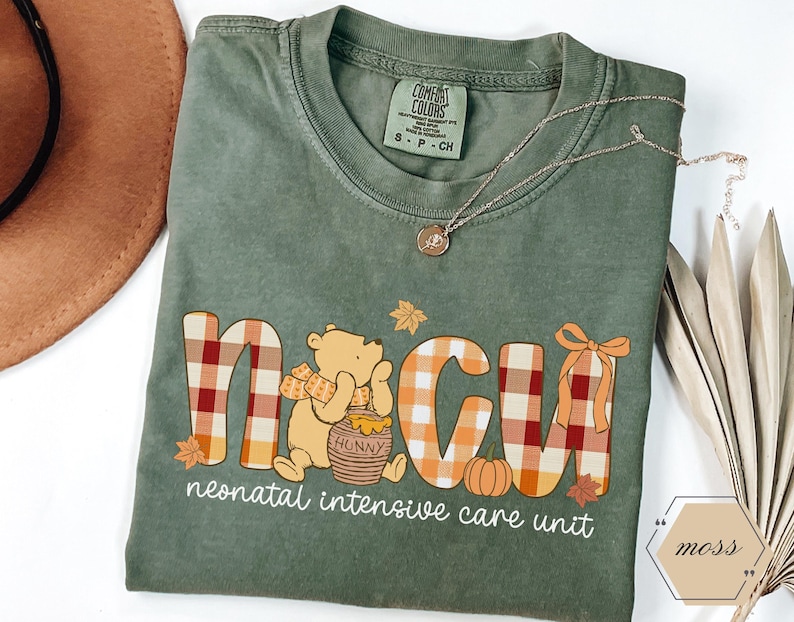 May include: Green Comfort Colors t-shirt with a Winnie the Pooh-themed design. The design includes the letters "NICU" in a plaid pattern, a bear, a pumpkin, and the words "neonatal intensive care unit". A gold necklace is draped across the shirt.