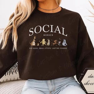 Pu&ograve; includere: Felpa color cioccolato fondente con la scritta bianca "SOCIAL WORKER". Sotto il testo ci sono personaggi dei cartoni animati. La felpa ha anche la scritta "BIG HUGS, SMALL STEPS, LASTING CHANGE".