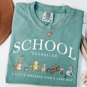 May include: A light green t-shirt featuring the white text SCHOOL COUNSELOR and the phrase A LITTLE KINDNESS GOES A LONG WAY. Below the text are illustrations of various cartoon characters including a kangaroo, bear, rabbit, tiger, pig, and donkey.