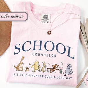 May include: A light pink t-shirt featuring the dark blue text SCHOOL COUNSELOR and the phrase A LITTLE KINDNESS GOES A LONG WAY. Below the text are illustrations of Winnie the Pooh characters including Roo, Kanga, Pooh, Rabbit, Tigger, Piglet, and Eeyore.