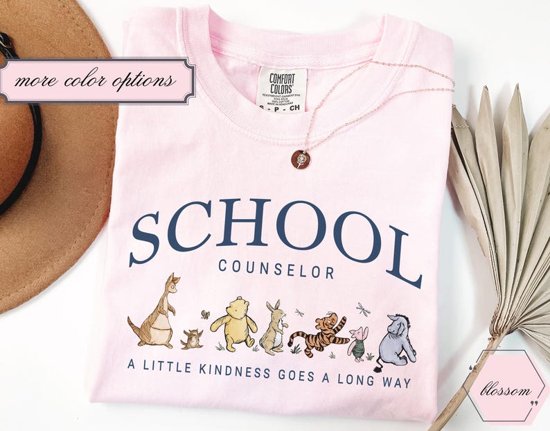 May include: A light pink t-shirt featuring the dark blue text SCHOOL COUNSELOR and the phrase A LITTLE KINDNESS GOES A LONG WAY. Below the text are illustrations of Winnie the Pooh characters including Roo, Kanga, Pooh, Rabbit, Tigger, Piglet, and Eeyore.