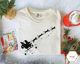 May include: Ivory-colored t-shirt featuring a black graphic of Santa Claus in a sleigh pulled by cats, with stars and dots. The shirt has a crew neck and a tag that reads "Comfort Colors". A holiday card and a cup of coffee are in the background.