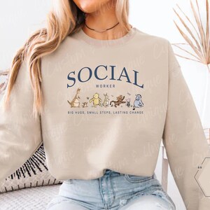 Pu&ograve; includere: Una felpa beige con la scritta "SOCIAL WORKER" in blu navy, arcuata sul petto. Sotto, personaggi dei cartoni animati. Il testo "BIG HUGS, SMALL STEPS, LASTING CHANGE" &egrave; stampato sotto.