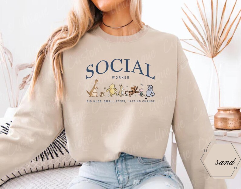 Pu&ograve; includere: Una felpa beige con la scritta "SOCIAL WORKER" in blu navy, arcuata sul petto. Sotto, personaggi dei cartoni animati. Il testo "BIG HUGS, SMALL STEPS, LASTING CHANGE" &egrave; stampato sotto.