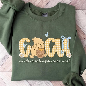 May include: A military green sweatshirt featuring the letters "CCU" in a yellow gingham pattern, a Winnie the Pooh graphic, and the words "cardiac intensive care unit". Butterflies and flowers are also included.