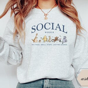 Pu&ograve; includere: Una felpa grigio chiaro con la scritta "SOCIAL WORKER" in blu navy arcuata nella parte superiore. Sotto il testo c'&egrave; una fila di personaggi dei cartoni animati. Il testo "BIG HUGS, SMALL STEPS, LASTING CHANGE" &egrave; sotto.