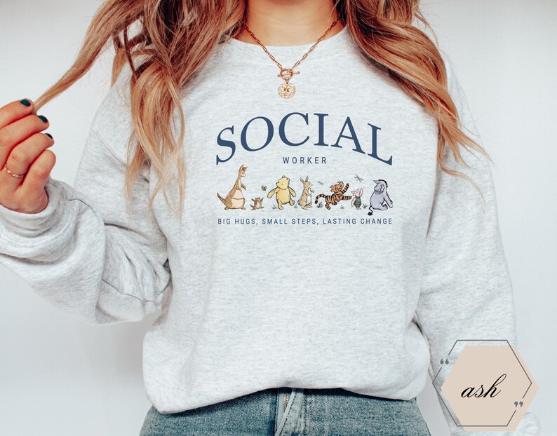 Pu&ograve; includere: Una felpa grigio chiaro con la scritta "SOCIAL WORKER" in blu navy arcuata nella parte superiore. Sotto il testo c'&egrave; una fila di personaggi dei cartoni animati. Il testo "BIG HUGS, SMALL STEPS, LASTING CHANGE" &egrave; sotto.
