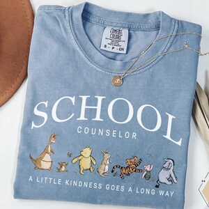 May include: A blue short-sleeved t-shirt with white text that reads SCHOOL COUNSELOR and A LITTLE KINDNESS GOES A LONG WAY. The shirt features a graphic of various cartoon animals including a bear, a tiger, a donkey, and a kangaroo.