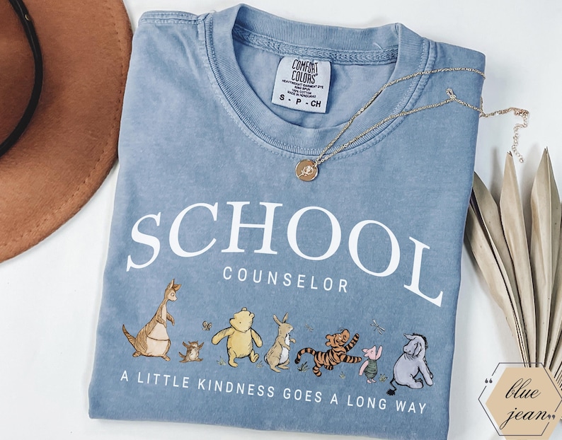 May include: A blue short-sleeved t-shirt with white text that reads SCHOOL COUNSELOR and A LITTLE KINDNESS GOES A LONG WAY. The shirt features a graphic of various cartoon animals including a bear, a tiger, a donkey, and a kangaroo.