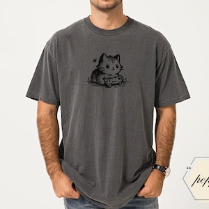 May include: Dark gray t-shirt featuring a black and white illustration of a cat playing a video game. The cat is holding a game controller and is surrounded by a star and grass. The shirt is a relaxed fit.