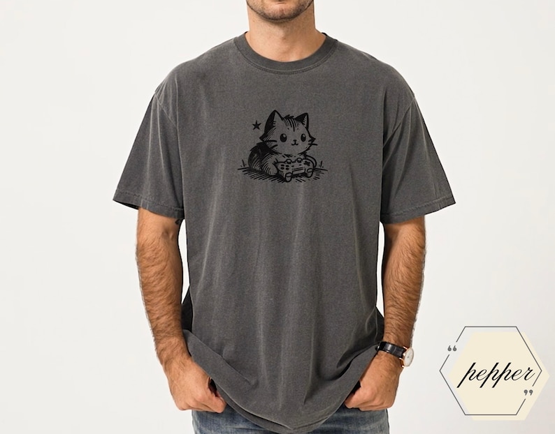 May include: Dark gray t-shirt featuring a black and white illustration of a cat playing a video game. The cat is holding a game controller and is surrounded by a star and grass. The shirt is a relaxed fit.