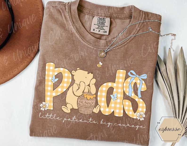 May include: Brown t-shirt with the word "Pods" in yellow gingham print, featuring a cartoon bear and honey pot. The text "little patients, big courage" is below. A silver necklace and a small bee graphic are also visible.