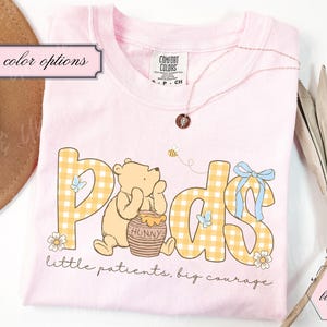 May include: A light pink t-shirt featuring a cartoon bear with the word "Peds" in yellow gingham letters. The shirt includes the phrase "little patients, big courage" and a small bee graphic. A necklace and a hat are visible.