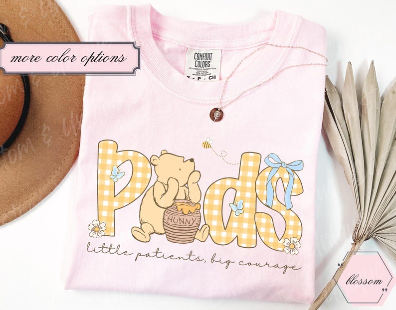 May include: A light pink t-shirt featuring a cartoon bear with the word "Peds" in yellow gingham letters. The shirt includes the phrase "little patients, big courage" and a small bee graphic. A necklace and a hat are visible.