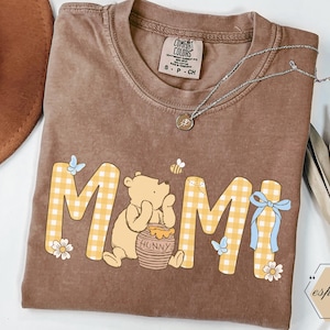 以下が含まれることがあります： 黄色のギンガムチェック柄で「MAMI」の文字が入った茶色のコンフォートカラーズTシャツ。くまのプーさんのキャラクター、ハチミツの壺、蝶、蜂が描かれています。ペンダント付きのシルバーネックレスも写っています。
