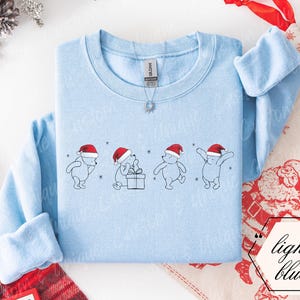 Könnte beinhalten: Ein hellblaues Sweatshirt mit Rundhalsausschnitt und einem zentralen Aufdruck von vier Winnie Puuh-Figuren mit roten Weihnachtsmützen. Die Figuren sind in verschiedenen Posen dargestellt, eine davon sitzt neben einem Geschenk. Mit kleinen Schneeflocken.