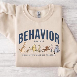May include: A sand-colored sweatshirt with the words "BEHAVIOR ANALYST" in navy blue arched across the top. Below the text are cartoon characters. The phrase "SMALL STEPS MAKE BIG PROGRESS" is printed below the characters.