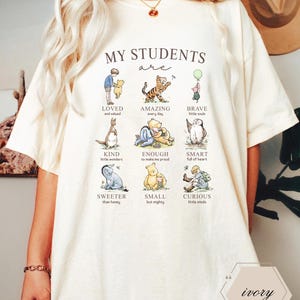 May include: Ivory-colored t-shirt featuring illustrations of Winnie the Pooh characters with positive adjectives. The text "MY STUDENTS are" is above the images. The shirt has a relaxed fit and short sleeves, perfect for casual wear.