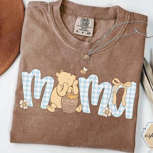 May include: Brown Comfort Colors t-shirt with a Winnie the Pooh graphic. The word "mama" is in blue gingham print with a bear, honey pot, bee, and flowers. A silver necklace with a pendant is on the shirt.