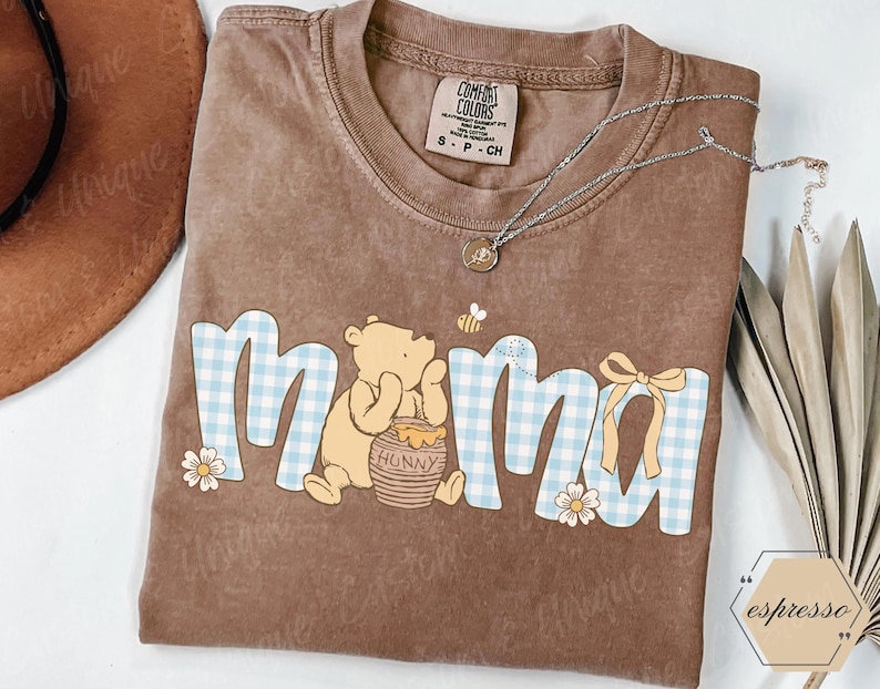May include: Brown Comfort Colors t-shirt with a Winnie the Pooh graphic. The word "mama" is in blue gingham print with a bear, honey pot, bee, and flowers. A silver necklace with a pendant is on the shirt.