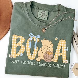 May include: Green t-shirt featuring the letters "BOA" in yellow gingham, a Winnie the Pooh graphic, and the text "BOARD CERTIFIED BEHAVIOR ANALYST". A gold necklace and a brown hat are also visible.