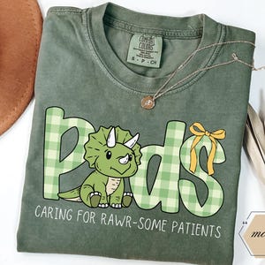 May include: A sage green t-shirt featuring the word "Pads" in green plaid letters and a cartoon triceratops. The text "CARING FOR RAWR-SOME PATIENTS" is below the dinosaur. A gold necklace is draped across the shirt.