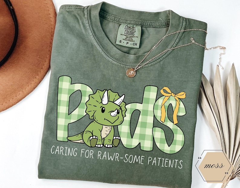 May include: A sage green t-shirt featuring the word "Pads" in green plaid letters and a cartoon triceratops. The text "CARING FOR RAWR-SOME PATIENTS" is below the dinosaur. A gold necklace is draped across the shirt.