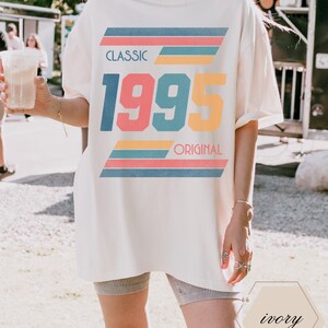 May include: A white oversized t-shirt with a retro graphic design featuring the text "CLASSIC 1995 ORIGINAL" in colorful block letters with stripes.