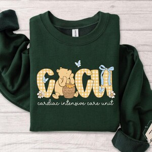 May include: Dark green sweatshirt with the letters "CICU" in a yellow gingham pattern, featuring a cartoon bear and the words "cardiac intensive care unit". The design includes butterflies, flowers, and a honey pot.