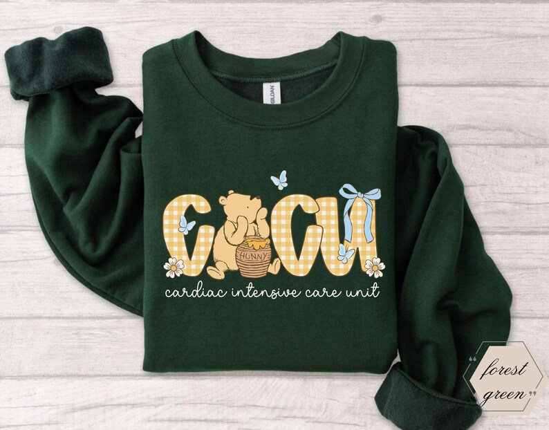 May include: Dark green sweatshirt with the letters "CICU" in a yellow gingham pattern, featuring a cartoon bear and the words "cardiac intensive care unit". The design includes butterflies, flowers, and a honey pot.