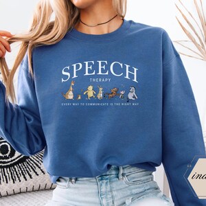 May include: Blue sweatshirt with the words "SPEECH THERAPY" in white arched text above a line of cartoon characters. Below the text is the phrase "EVERY WAY TO COMMUNICATE IS THE RIGHT WAY".