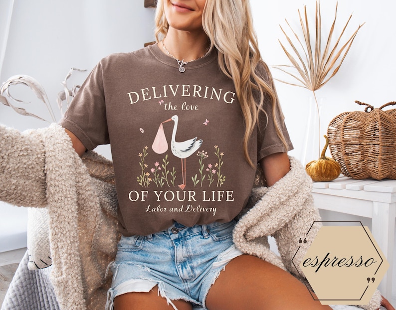 Op de afbeelding: Een bruin t-shirt met een afbeelding van een ooievaar die een baby draagt en de tekst "Delivering the love of your life Labor and Delivery".