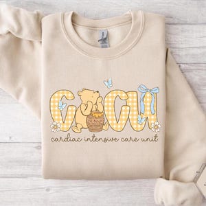 May include: A sand-coloured sweatshirt with the letters "GCIU" in a yellow gingham pattern, featuring a cartoon bear and the words "cardiac intensive care unit". The design includes butterflies and flowers.