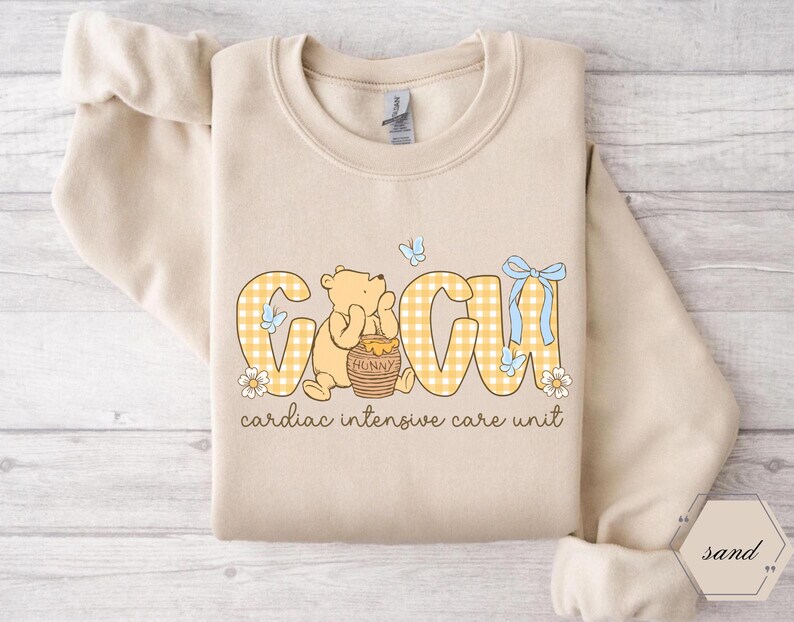May include: A sand-coloured sweatshirt with the letters "GCIU" in a yellow gingham pattern, featuring a cartoon bear and the words "cardiac intensive care unit". The design includes butterflies and flowers.