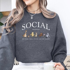 Pu&ograve; includere: Felpa grigio scuro con la scritta bianca "SOCIAL WORKER" a forma di arco. Sotto il testo ci sono personaggi dei cartoni animati. La frase "BIG HUGS, SMALL STEPS, LASTING CHANGE" &egrave; stampata sotto i personaggi.