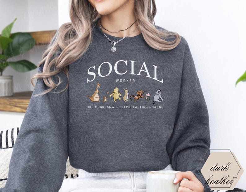 Pu&ograve; includere: Felpa grigio scuro con la scritta bianca "SOCIAL WORKER" a forma di arco. Sotto il testo ci sono personaggi dei cartoni animati. La frase "BIG HUGS, SMALL STEPS, LASTING CHANGE" &egrave; stampata sotto i personaggi.