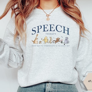 May include: A light gray sweatshirt with the words "SPEECH THERAPY" in navy blue arched across the top. Below the text is a line of cartoon characters. The phrase "EVERY WAY TO COMMUNICATE IS THE RIGHT WAY" is printed below the characters.