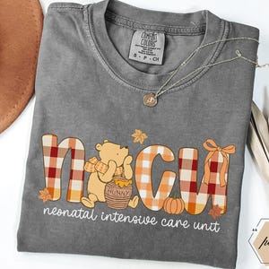 May include: A gray Comfort Colors t-shirt with a Winnie the Pooh-themed design. The design spells out NICU with a plaid pattern and fall accents. The words "neonatal intensive care unit" are printed below the graphic.