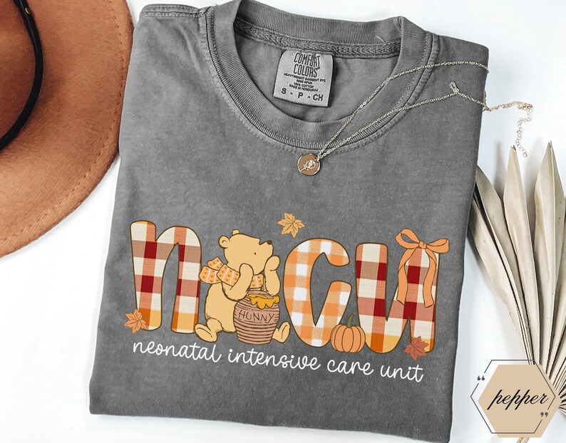 May include: A gray Comfort Colors t-shirt with a Winnie the Pooh-themed design. The design spells out NICU with a plaid pattern and fall accents. The words "neonatal intensive care unit" are printed below the graphic.