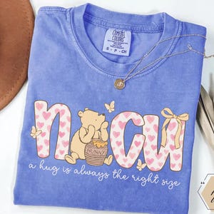 May include: A blue t-shirt featuring the word "NCU" in a playful font filled with pink hearts. A cartoon bear sits beside a honey pot. The text "a hug is always the right size" is below the graphic.