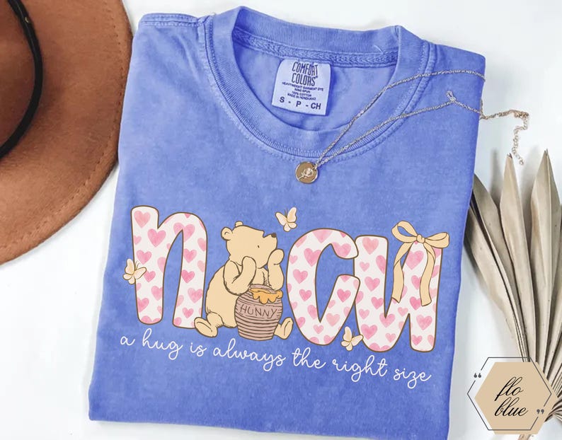 May include: A blue t-shirt featuring the word "NCU" in a playful font filled with pink hearts. A cartoon bear sits beside a honey pot. The text "a hug is always the right size" is below the graphic.