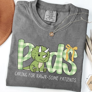 May include: A grey t-shirt featuring the word "Pods" in green plaid lettering and a cartoon triceratops. The text "CARING FOR RAWR-SOME PATIENTS" is below. A gold necklace and a bow are also visible on the shirt.