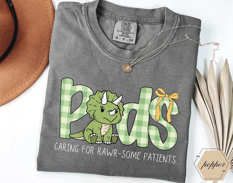 May include: A grey t-shirt featuring the word "Pods" in green plaid lettering and a cartoon triceratops. The text "CARING FOR RAWR-SOME PATIENTS" is below. A gold necklace and a bow are also visible on the shirt.