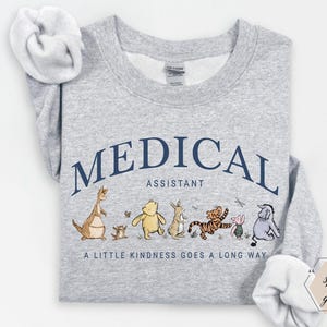 Op de afbeelding: Een lichtgrijs sweatshirt met de marineblauwe tekst "MEDICAL ASSISTANT" in een boog. Daaronder een rij cartoonfiguren. De zin "A LITTLE KINDNESS GOES A LONG WAY" is eronder gedrukt.
