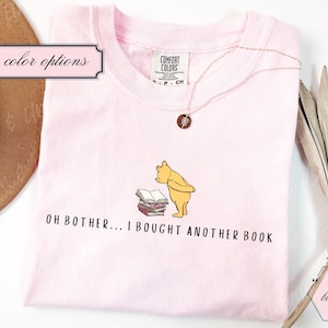 May include: Light pink t-shirt featuring a cartoon illustration of Winnie the Pooh looking at a stack of books. The text "OH BOTHER... I BOUGHT ANOTHER BOOK" is printed below the image. A rose gold necklace is draped on the shirt.