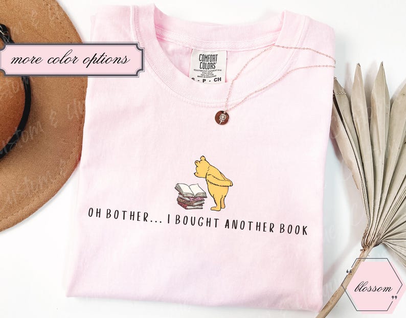 May include: Light pink t-shirt featuring a cartoon illustration of Winnie the Pooh looking at a stack of books. The text "OH BOTHER... I BOUGHT ANOTHER BOOK" is printed below the image. A rose gold necklace is draped on the shirt.