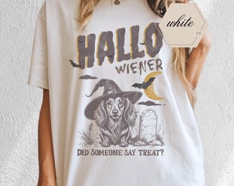 Puede incluir: Camiseta blanca con un diseño de Halloween. El diseño presenta las palabras "Hallo Wiener" en una fuente desgastada, un perro salchicha con un sombrero de bruja, una lápida, un murciélago y una luna creciente. También incluye la frase "Did Someone Say Treat?".